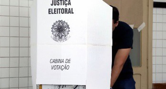 homem atrás de cabine de votação com o símbolo da justiça eleitoral