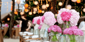 Evento com flores brancas e rosas em vasos de vidro em cima de uma mesa de madeira; guardanapos brancos com talheres sob a mesa também; fundo com luzes desfocadas