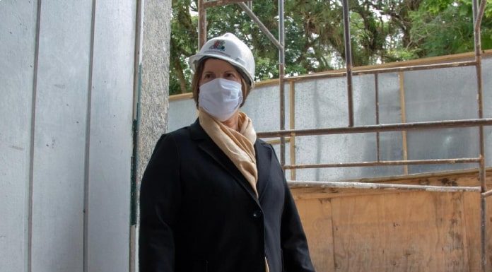 prefeita adeliana usando máscara e capacete na porta do teatro em obras; ela usa um sobretudo e cachecol