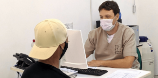 Homem com uniforme da Prefeitura de Biguaçu, sentado de um lado da mesa e mexendo no computador, e um segundo homem, paciente, de costas esperando ser marcada a sua consulta - Consultas de rotina voltam a ser agendadas nos postos de saúde de Biguaçu