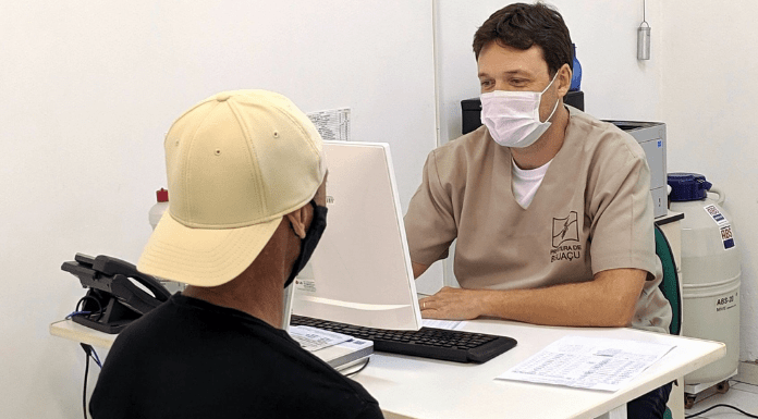 Homem com uniforme da Prefeitura de Biguaçu, sentado de um lado da mesa e mexendo no computador, e um segundo homem, paciente, de costas esperando ser marcada a sua consulta - Consultas de rotina voltam a ser agendadas nos postos de saúde de Biguaçu