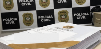 Inquérito policial finalizado em cima de uma mesa na delegacia, atrás o painel da Polícia Civil de Santa Catarina