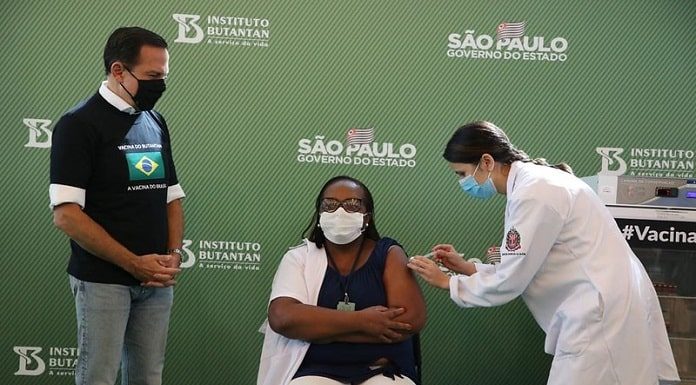 monica, primeira vacinada contra coronavírus, sentada recebe a dose no braço dada pela enfermeira, observadas pelo governador de são paulo, joão dória, todos usam máscaras