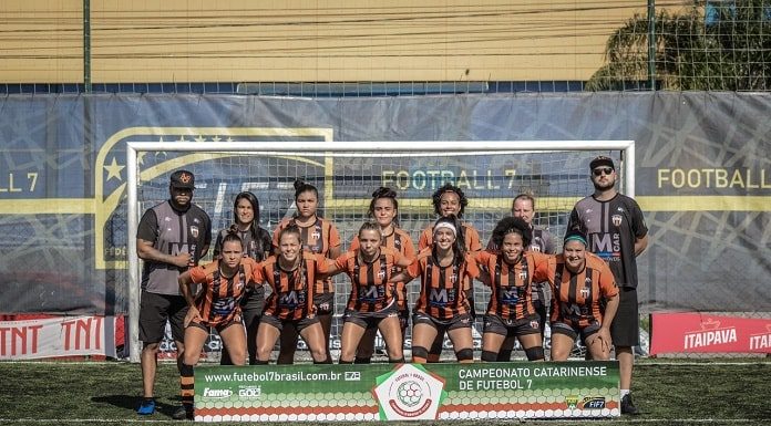 mulheres postadas para foto do time do atlético catarinense; todas sorridentes na frente do gol e com placa no chão à frente