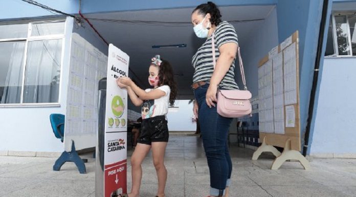 Menina de máscara, acompanhada de uma mulher adulta também de máscara, pega álcool em gel em um toten em frente a escola. Máscaras e álcool em gel são parte dos EPIs que serão distribuídos nas escola estaduais de SC.