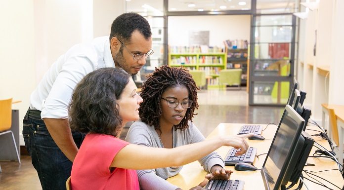 Uma mulher sentada aponta para a tela do computador enquanto outra mulher, sentada ao seu lado, digita no teclado. Atrás delas está um homem acompanhando o trabalho. A imagem ilustra o estágio de estudantes em empresas, como as de tecnologia, que abriram vagas em SC.