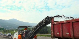 maquina recortando asfalto da via expressa sul e jogando detrito em caçamba de caminhão; operários ao lado; trânsito ao fundo