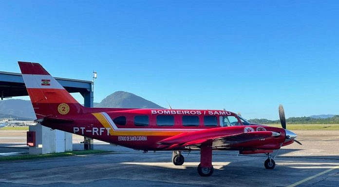 Na foto observa-se um céu azul e um avião arcanjo do corpo de bombeiros parado na pista. O arcanjo é vermelho com detalhes em amarelo e identificação em branco. O veículo fará a transferência de pacientes de SC para o Espírito Santo.