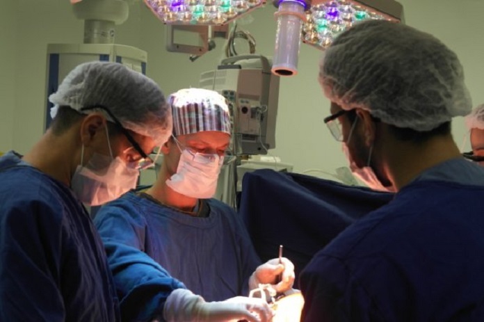 Três profissionais da saúde com roupas azuis, máscaras e proteção nos olhos e cabelos se concentram em um procedimento cirúrgico. As cirurgias eletivas em Santa Catarina estão suspensas na rede privada até o fim de março.