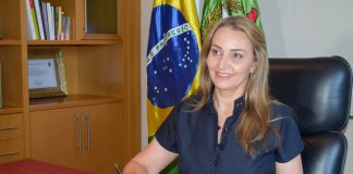 daniela reinher sorridente com caneta na mão sobre caderno sentada em mesa de escritório