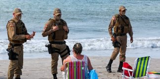 dois policiais usando máscara conversam com mulher sentada em cadeira de praia na beira do mar; outro policial de máscara ao lado - serviços que podem funcionar em sc no fim de semana