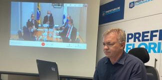 gean loureiro sentado em ponta de mesa com notebook à frente e painel com reunião virtual ao fundo