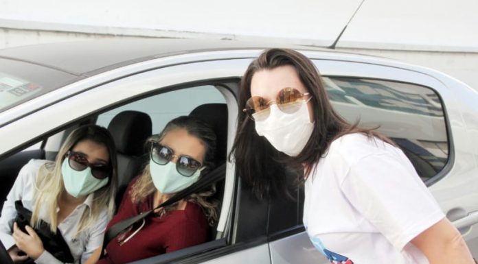 três mulheres usando máscara e óculos escuros posam para foto; duas estão dentro do carro e uma próxima, do lado de fora, em drive-thru de vacinação