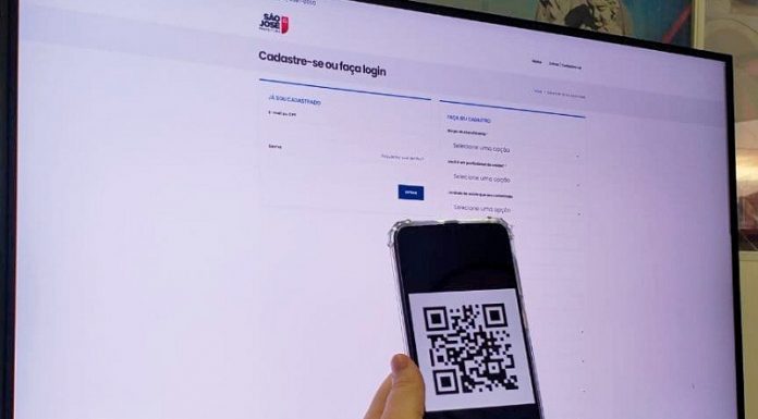 tela de um computador em página branca com logo de são josé na parte de cima, é o site de cadastro para vacinas do município, a frente da tela uma mão segura um celular com um qr code aberto.