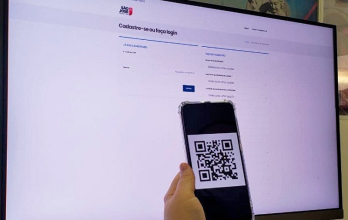 tela de um computador em página branca com logo de são josé na parte de cima, é o site de cadastro para vacinas do município, a frente da tela uma mão segura um celular com um qr code aberto.