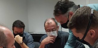 homens reunidos em volta de uma mesa, usando máscaras, olham e apontam para papeis com projetos