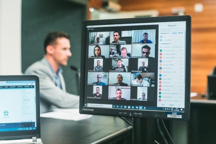 O foco da foto é em uma tela de computador que mostra 15 quadrados com o vídeo de participantes da audiência pública sobre o funcionamento das arenas de atividades esportivas de SC. Ao fundo o deputado que solicitou o encontro.