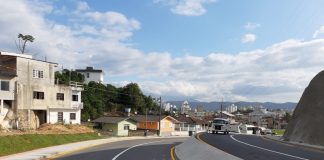 A foto mostras a avenida das torres em Palhoça, com asfalta novo e pista dupla. O céu é azul e ao fundo é possível ver algumas residencias.