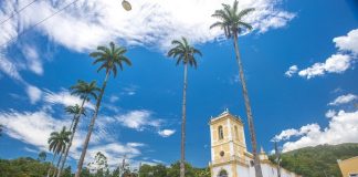 A foto com ângulo de baixo para cima mostra o céu azul e ensolarado com nuvens brancas, há árvores altas e é possível ver construções históricas. A prefeitura de palhoça organiza um concurso fotográfico em seu aniversário de emancipação.