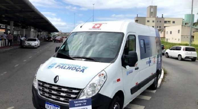 Ambulância branca com identificação em azul da prefeitura de Palhoça. O veículo está parado em frente a estação Palhoça, onde ocorre a vacinação do município.