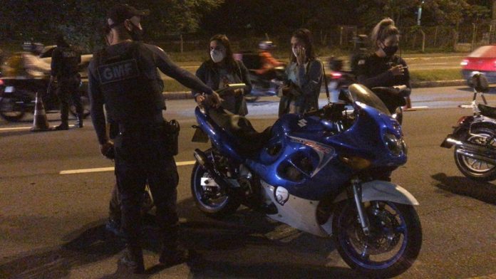 A foto em período noturno mostra uma moto parada com dois guardas municipais e duas pessoas conversando com eles, todo usam máscaras. Ao fundo uma movimentação de moto e carros. A GMF passa a utilizar um aparelho para identificas as motos em alta velocidade e emissão de ruídos