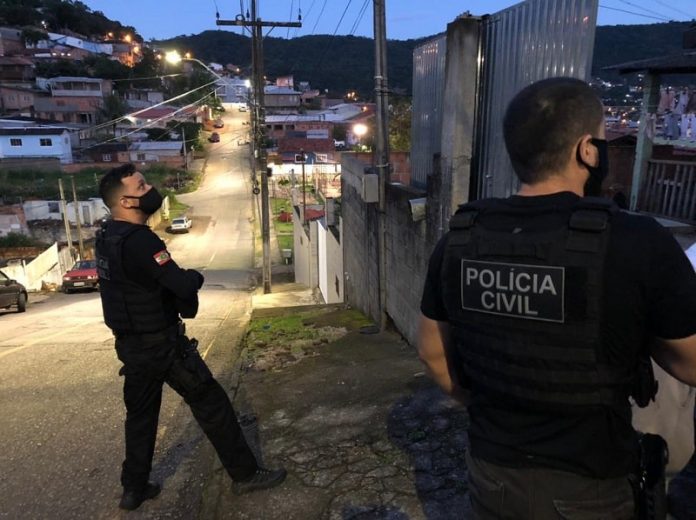 dois policiais civis olham para residência em ladeira com muitas casas ao fundo no início da 3ª fase da operação héracles