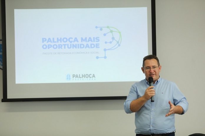 O prefeito Eduardo Freccia é branco, tem cabelo escuros e curtos, usa uma camisa azul clara e segura um microfone. Ao fundo, um telão mostra: Palhoça Mais Oportunidade, progrrama de retomada ecônomica e social