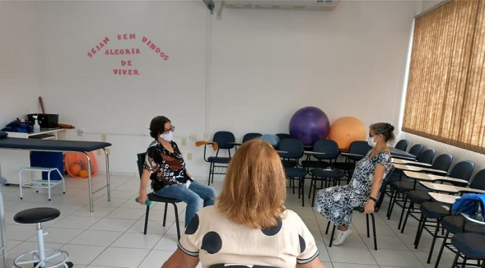 Em uma sala, aparece de costas uma senhora de cabelos claros sentada em uma cadeira com os braços abertos, a sua frente duas mulheres de máscara, sentadas uma de frente para a outra copiam o movimento com os braços. Palhoça oferece o tratamento de fisioterapia para pacientes que tiveram covid-19.