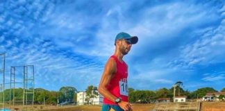 Um atleta de camiseta vermelha, tênis, boné e shorts azuis, além de óculos escuros corre em marcha atlética em uma pista de cor azul. O time de atletismo de São José conquistou vaga nos jogos sul-americanos