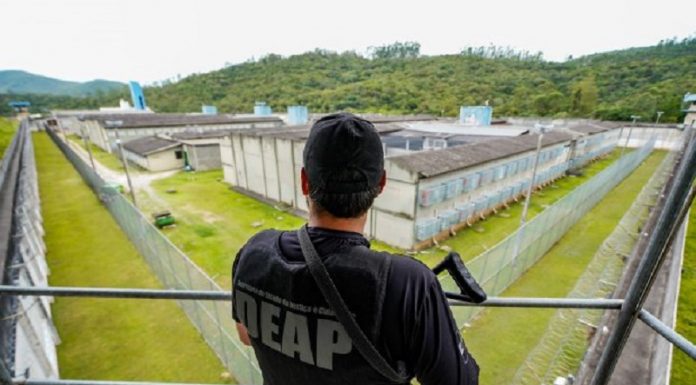 De costas, uma pessoa aparece de boné, camiseta e colete pretos, ele carrega um armamento e observa uma área que parece ser um presídio. Nesta semana, o governo convocou 213 policias penais