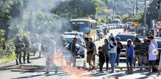 Na imagem um grupo de pessoas protesta, há um ponto de fogo em toras na pista e uma fila de carros. Os moradores do Morro das Pedras protestaram por resoluções para o problema de destruição de imóveis na beira da praia