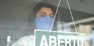 mulher com roupa branca e máscara azul colocando uma placa de aberto na porta do estabelecimento