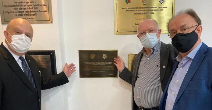 presidentes de avaí e figueirense e mais um homem, todos de máscara, olha para foto em frente à placa de homenagem dourada