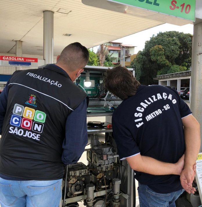 Duas pessoas de cabelo curto estão de costas olhando para uma bomba de um posto de combustível. Um veste um colete escuro do Procon de São José e outro veste uma camiseta preta em que se lê fiscalização imetro/sc. A operação transparência lacrou bombas irregulares durante a fiscalização em postos de combustíveis de são josé.