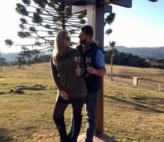 Roberta Zilli e Bruno Buske olham um para o outro em paisagem serrana segurando taça de vinho