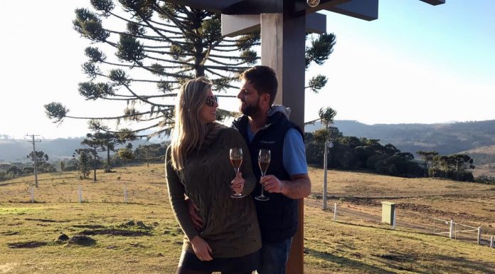 Roberta Zilli e Bruno Buske olham um para o outro em paisagem serrana segurando taça de vinho