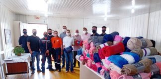 Um grupo de pessoas aparece lado a lado posando para a foto, ao lado, no canto direito, uma mesa com os cobertores doados para famílias da Frei Damiçao, em Palhoça.