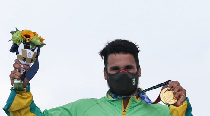 Ítalo Ferreira tem cabelos escuros e curtos, usa uma máscara preta e um agasalho verde e amarelo do Brasil, ele segura com a mão a medalha de ouro, conquistada no surfe, enroscada no pescoço e na outra mão levanta as flores entregues junto com a medalha nas olimpíadas de tóquio.