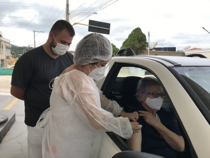 Uma profissional de saúde com vestimenta brancas aplica a vacina da covid-19 em um senhor dentro de um veículo, em Nova Trento, ao lado do carro, do lado de fora, um rapaz observa. O município de Nova Trento abriu nesta semana o cadastro para xepa da vacina contra a covid-19.