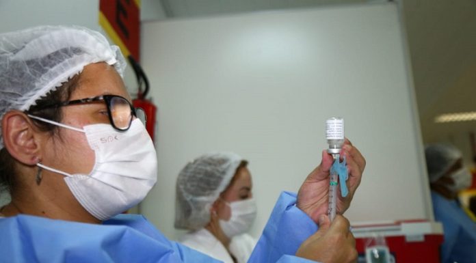 Uma profissional de saúde com vestimentas azuis e máscara e touca branca, além de ´pculos de grau, segura a sua frente um frasco e uma seringa de vacina. A foto ilustra a notícia da liberação do uso de xepa da vacina para adolescentes comd eficiências ou comorbidades em SC.