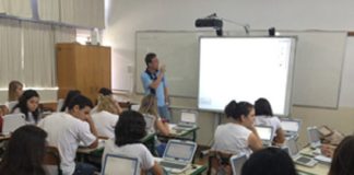 Uma sala de aula com um professor a frente olhando para um telão e observado por estudantes nas carteiras com computadores. O novo ensino médio em SC entra em vigor em 2022 nas escolas estaduais.