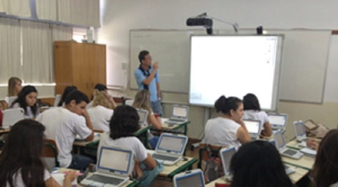 Uma sala de aula com um professor a frente olhando para um telão e observado por estudantes nas carteiras com computadores. O novo ensino médio em SC entra em vigor em 2022 nas escolas estaduais.