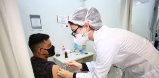 Um homem de cabelo escuro veste camiseta e máscara pretas, ele está sentado e recebe a vacina contra a covid no braço, aplicada por uma profissional da saúde com vestimentas brancas. Neste sábado, Florianópolis vai ampliar a vacinação contra a Covid-19 para pessoas de 26 anos.