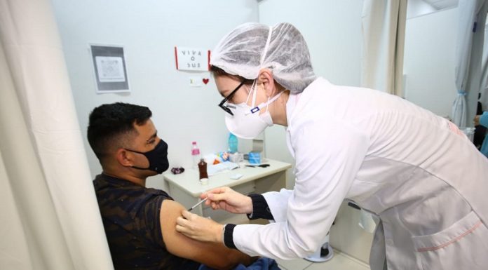 Um homem de cabelo escuro veste camiseta e máscara pretas, ele está sentado e recebe a vacina contra a covid no braço, aplicada por uma profissional da saúde com vestimentas brancas. Neste sábado, Florianópolis vai ampliar a vacinação contra a Covid-19 para pessoas de 26 anos.