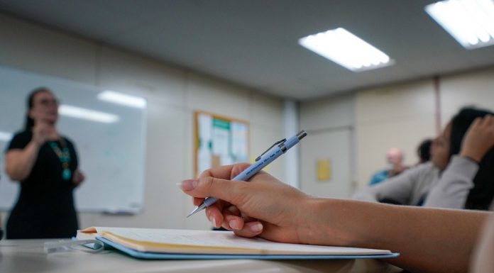 Edital publicado para concurso de ACTs em Santa Catarina - close em mão segurando caneta sobre caderno em sala de aula e professora em pé ao fundo dando aula