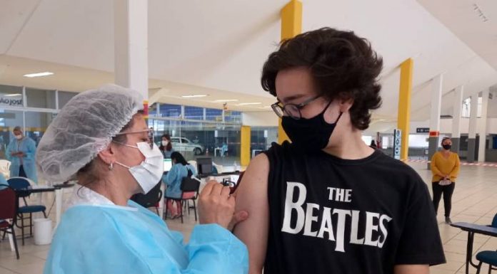 Vacinação contra Covid em adolescentes - rapaz usando camiseta dos beatles olha para enfermeira aplicando dose em seu braço