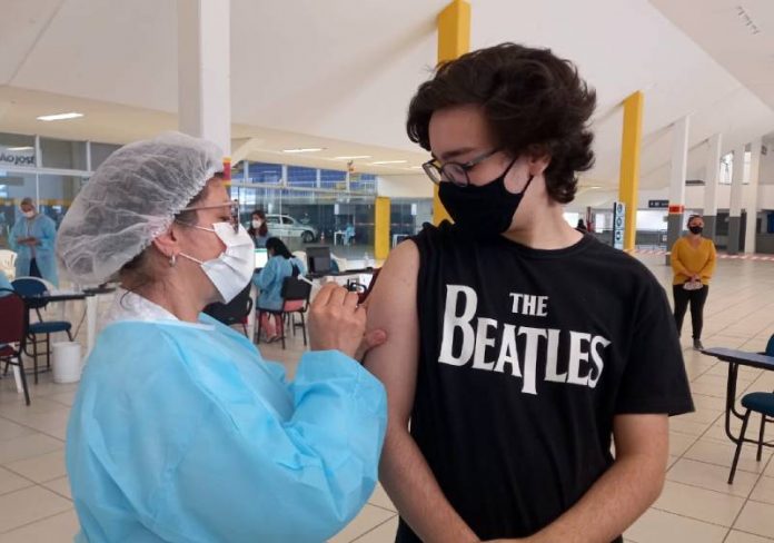 Vacinação contra Covid em adolescentes - rapaz usando camiseta dos beatles olha para enfermeira aplicando dose em seu braço