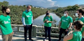 Seis pessoas de camisa verde e usando máscaras conversam em circulo, atrás deles o Rio Capivari. A foto ilustra a ação do programa Trato pelo Capivari, da Casan nos Ingleses, que realizou ação de conscientização com moradores do bairro.