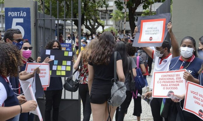 estudantes entrando em local de provam com pessoas no portão segurando cartazes e de máscara - Segundo dia do Enem tem provas de matemática e ciência da natureza