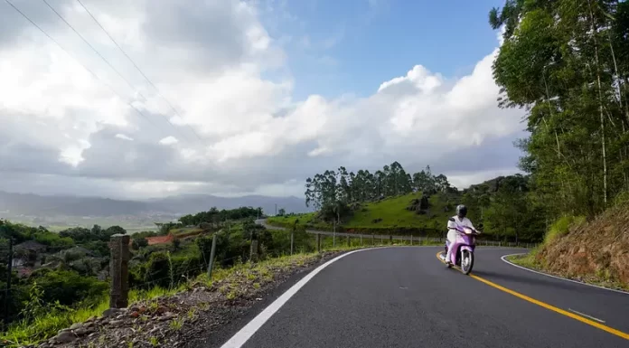 previsão do tempo | clima para amanhã em santa catarina: sol com algumas nuvens | mulher passando de biz em estrada do interior com grande pedaço de céu com nuvens e vale ao fundo